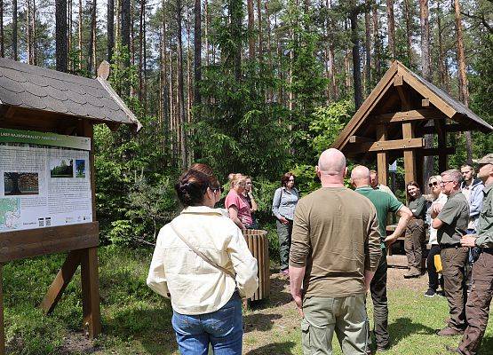 Osoby stojące przy wiacie i tablicy edukacyjnej na rowerowej szlaku "Pętla Przytarnia"