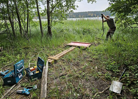 Na pierwszym planie widoczne narzędzia oraz leżąca na ziemi tablica informacyjna, za nią osoba ze szpadlem.