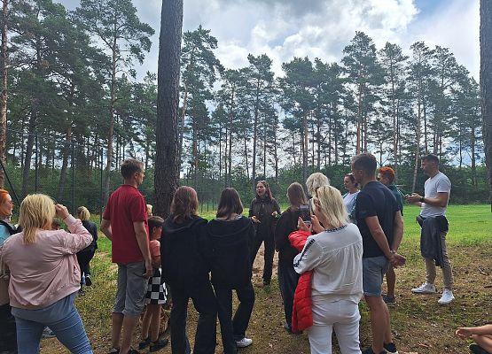 Grupa osób w leśnej polanie słucha prelegenta podczas wycieczki edukacyjnej na świeżym powietrzu.