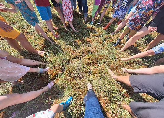 Krąg osób stojących boso lub w obuwiu na torfowisku, widoczny z góry, podczas plenerowego spotkania.
