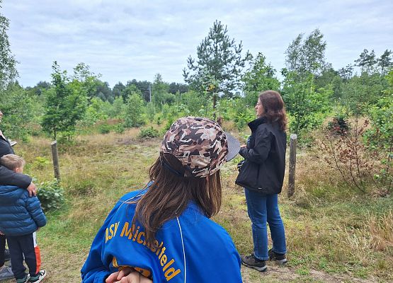 Uczestnicy wycieczki w lesie słuchają kobiety prowadzącej spotkanie, stojąc wśród młodych drzew i trawiastego terenu.