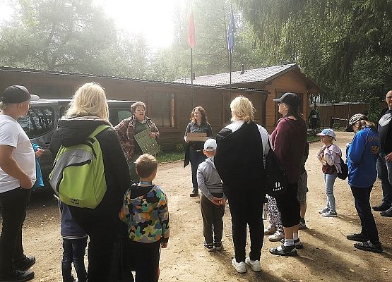 Przewodnicy prowadzą zajęcia dla grupy dzieci i dorosłych przy drewnianym budynku.