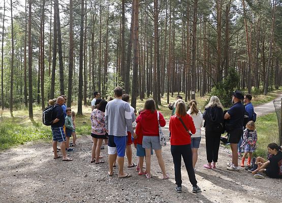 Grupa dorosłych i dzieci w lesie słucha przewodnika, który gestem uniesionej ręki prowadzi opowieść podczas spaceru edukacyjnego.