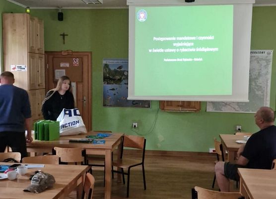 Zdjęcie przedstawia salę z zielonymi ścianami i drewnianą podłogą, w której kilka osób siedzi przy stołach. Na ekranie wyświetlana jest prezentacja w języku polskim.