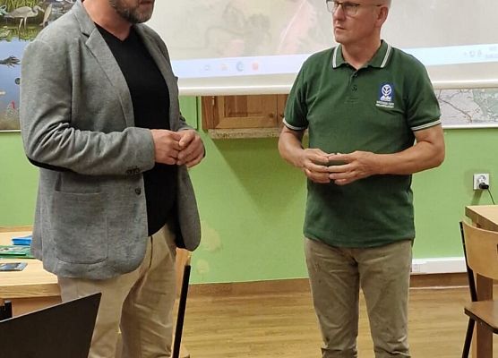 Dwaj mężczyźni rozmawiający na sali, w tle ekran multimedialny na którym prezentowany jest zdjęcie jeziora z lotu ptaka.