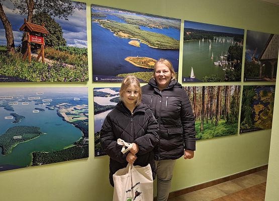 Dziecko z jedną dorosłą osoba pozujące do zdjęcia na tle ściany z obrazmi krajobrazowymi Wdzydzkiego Parku Krajobrazowego. Dziecko w dłoniach trzyma płócienną siatkę.