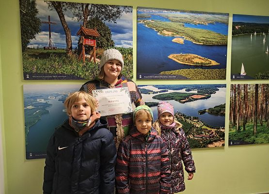 trójka dzieci i osoba dorosła pozujące do zdjęcia na tle ściany z obrazmi krajobrazowymi Wdzydzkiego Parku Krajobrazowego. Osoba dorosła trzyma w ręku dyplom.